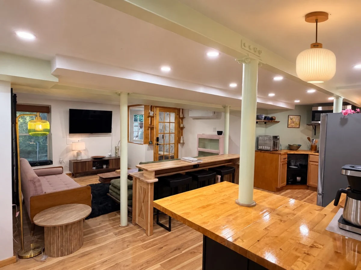 A New Life Spaces basement kitchen, finished: butcher-block counter spanning the original support columns, black stools, ribbed pendant lamp and oak floor.